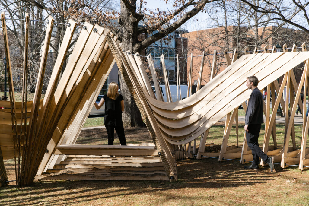 Two people standing with a structure made of wood that has been cut and pulled into long sheets. 