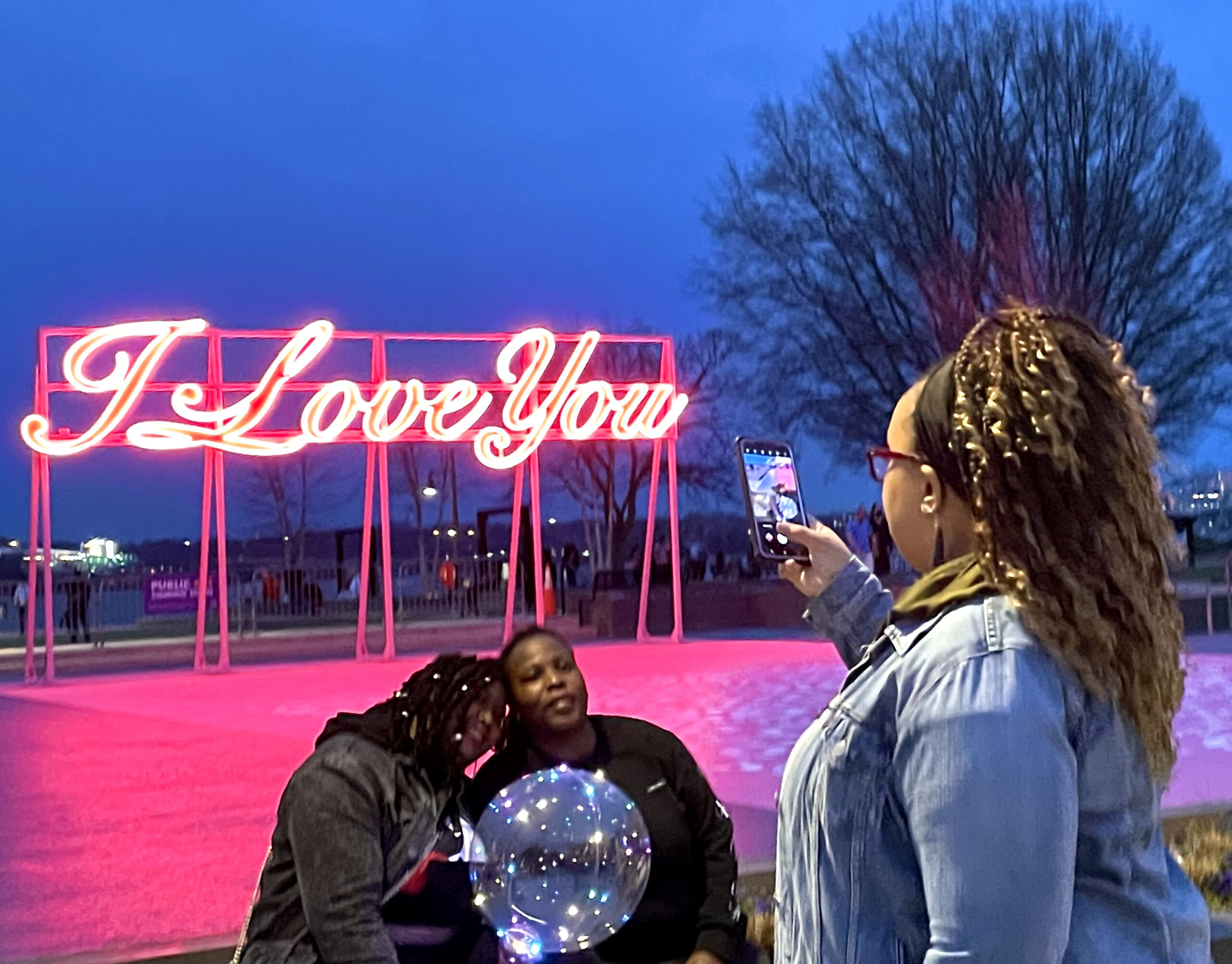 A woman with braids in the foreground taking a photograph of a couple standing in front of a neon pink sign that reads "I Love You"