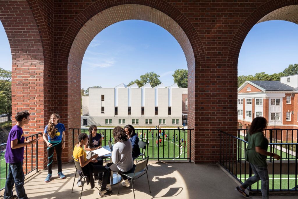 Students seated at a table with open archways overlooking a green courtyard in the background at a school for education.