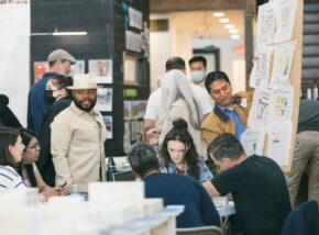 a group of people gathers in a space with designs pinned to the wall.